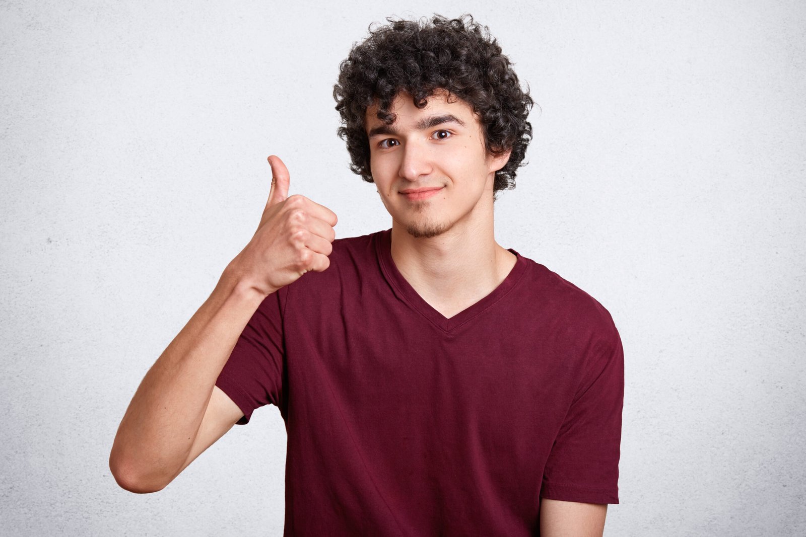 Handsome curly male with delighted expression, keeps thumb raised, shows his approval, poses against white concrete wall. Satisfied teenager with crisp hair shows ok sign. People and body language